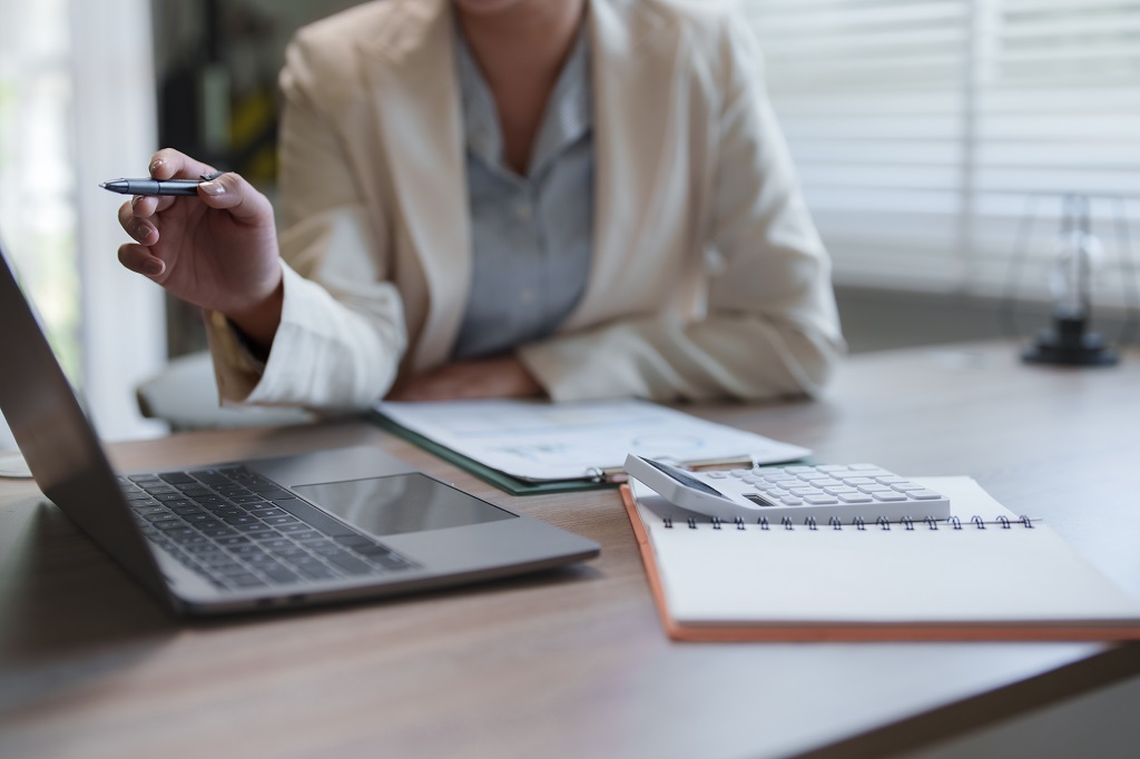Osoba za radnim stolom koristi laptop uz dokumenta, svesku i digitron